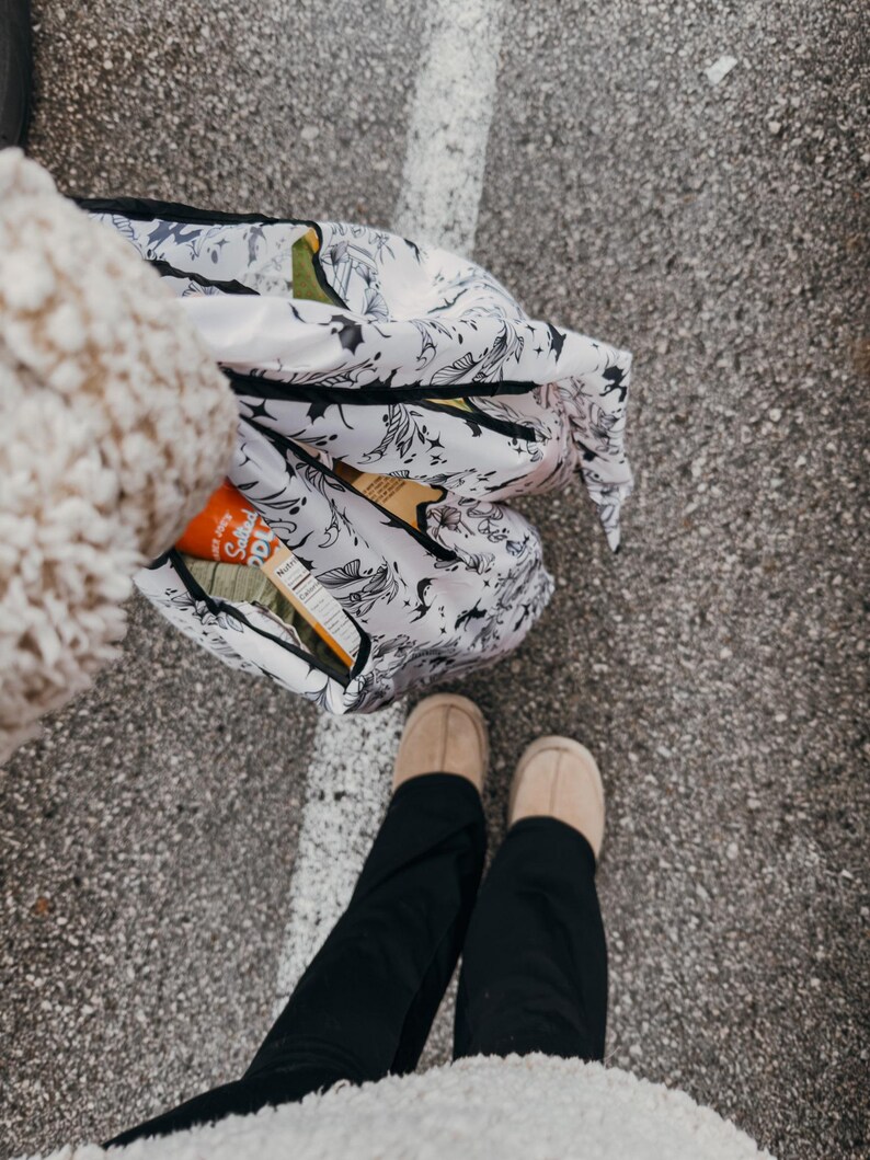 Peut inclure: Une personne portant un pantalon noir et un pull blanc duveteux tient un sac de courses r&eacute;utilisable blanc avec un motif floral noir et blanc. Le sac est rempli d'articles, dont une bo&icirc;te de craquelins. La personne se tient sur une surface d'asphalte grise.