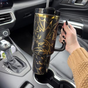 May include: A black and gold stainless steel tumbler with a floral and bat design. The tumbler has a handle and a straw. It is sitting in a cup holder in a car.