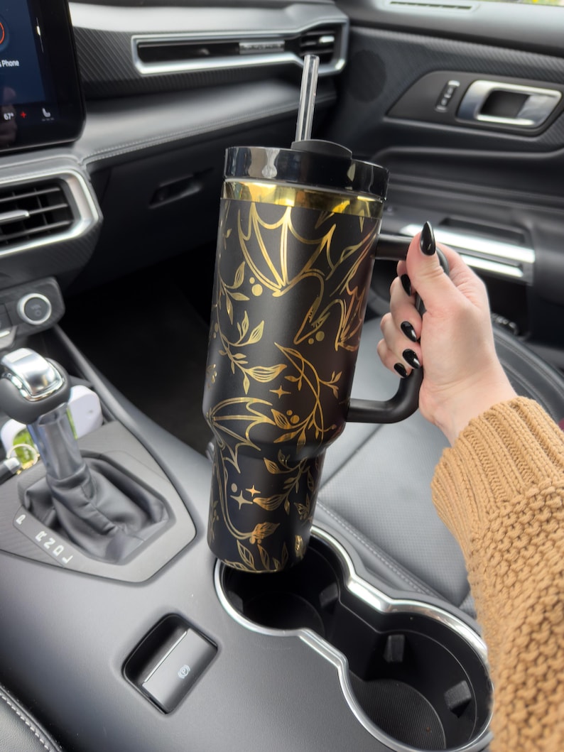 May include: A black and gold stainless steel tumbler with a floral and bat design. The tumbler has a handle and a straw. It is sitting in a cup holder in a car.