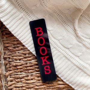 May include: A black bookmark with the word "BOOKS" in red letters. The bookmark is lying on a woven basket and a white knitted fabric.
