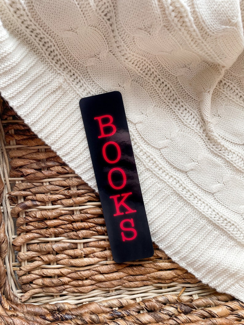 May include: A black bookmark with the word "BOOKS" in red letters. The bookmark is lying on a woven basket and a white knitted fabric.