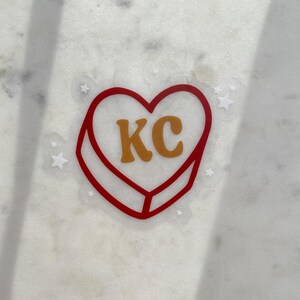 Puede incluir: Una pegatina transparente con un contorno rojo en forma de corazón y las letras "KC" en naranja. Pequeñas estrellas blancas rodean el corazón.