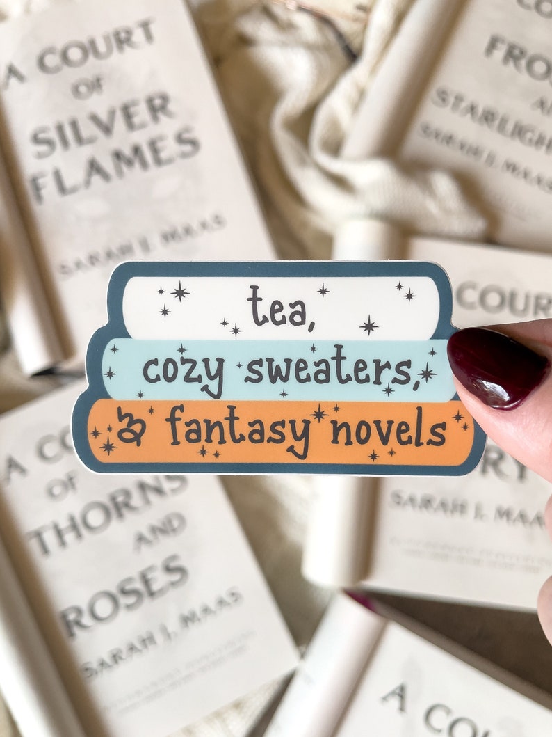May include: A sticker with the text "tea, cozy sweaters, & fantasy novels" in a blue, light blue, and orange color scheme. The sticker is decorated with small white stars.