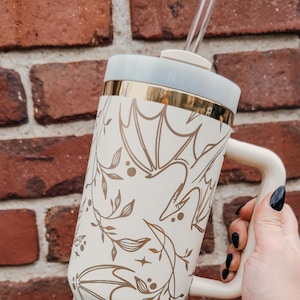 May include: A white travel mug with a gold rim and a floral pattern featuring gold leaves and stylized birds. The mug has a handle and a clear straw.