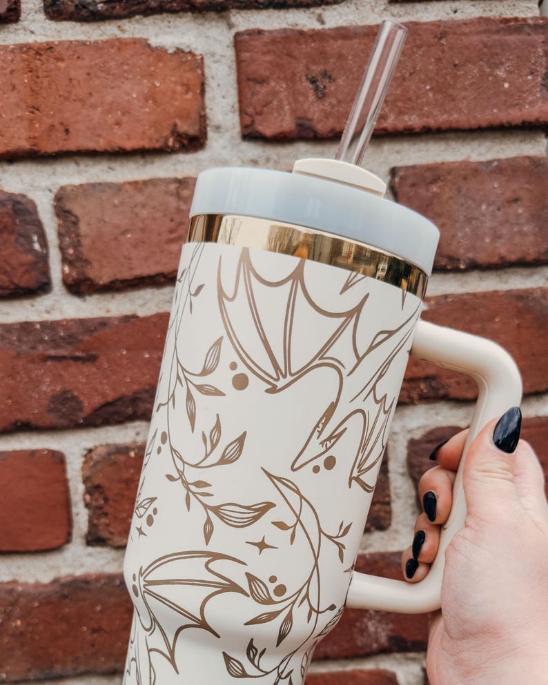 May include: A white travel mug with a gold rim and a floral pattern featuring gold leaves and stylized birds. The mug has a handle and a clear straw.