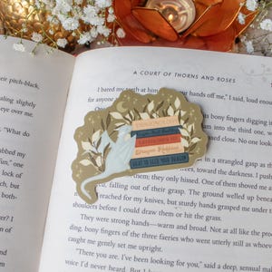 May include: A fantasy-themed bookmark with book titles including "Dragonology" and "What to Feed Your Dragon." The bookmark, with a light beige background and dragon design, sits on an open book. The design includes floral accents.