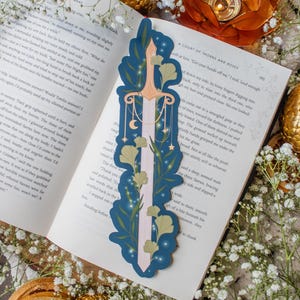 Puede incluir: Un marcapáginas decorativo con un diseño de espada, una hoja rosa y estrellas doradas, lunas y detalles florales. El marcapáginas está colocado en un libro con el texto "A COURT OF THORNS AND ROSES".