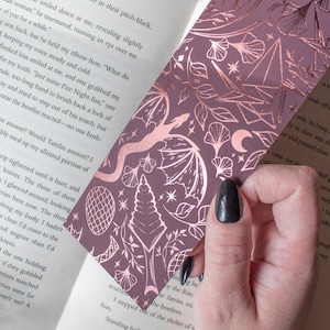 May include: A purple bookmark with a rose gold foil design featuring botanical and geometric shapes, including a dragon, leaves, and stars. The bookmark is placed in a book with text.