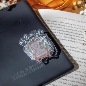May include: A clear sticker featuring a detailed illustration of a vintage armchair with the text "My Prince, My Love, My Woman". The sticker is placed on a black electronic device, with a book and decorative elements in the background.