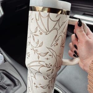 May include: A white and gold stainless steel tumbler with a floral and bat design. The tumbler has a lid and a straw. It is sitting in a car cup holder.