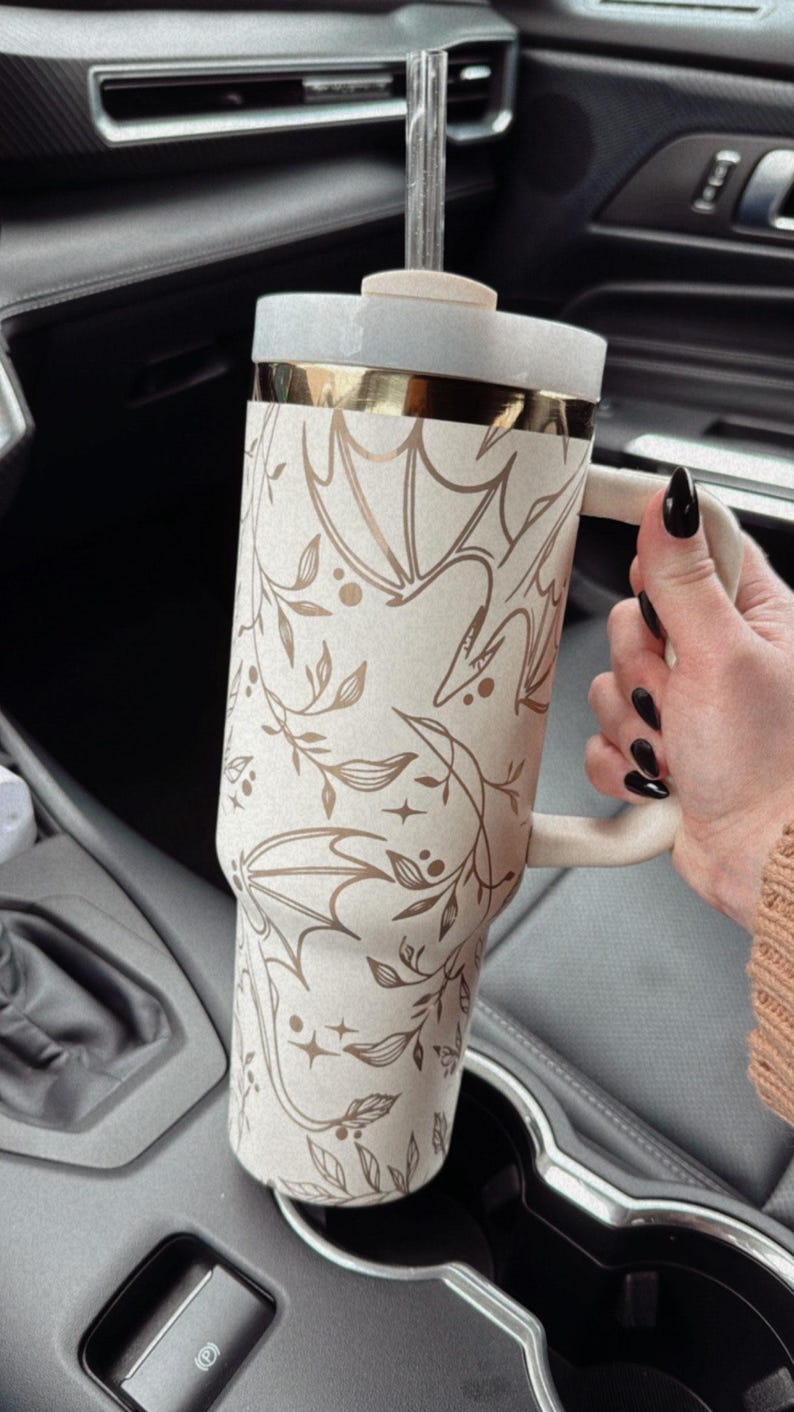 May include: A white and gold stainless steel tumbler with a floral and bat design. The tumbler has a lid and a straw. It is sitting in a car cup holder.