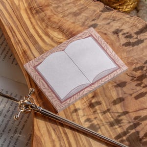 May include: A notepad with a book design, featuring a pink border with leaf accents, rests on a wooden surface. A gold-colored pen with a decorative top lies nearby. The notepad's pages are blank.