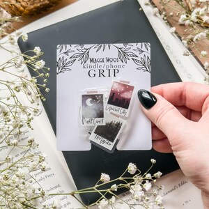May include: A white card with the text "MEAGGIE MOOS kindle/phone GRIP" and three small, rectangular grips with landscape designs. The grips are held over a black e-reader and an open book with white flowers.