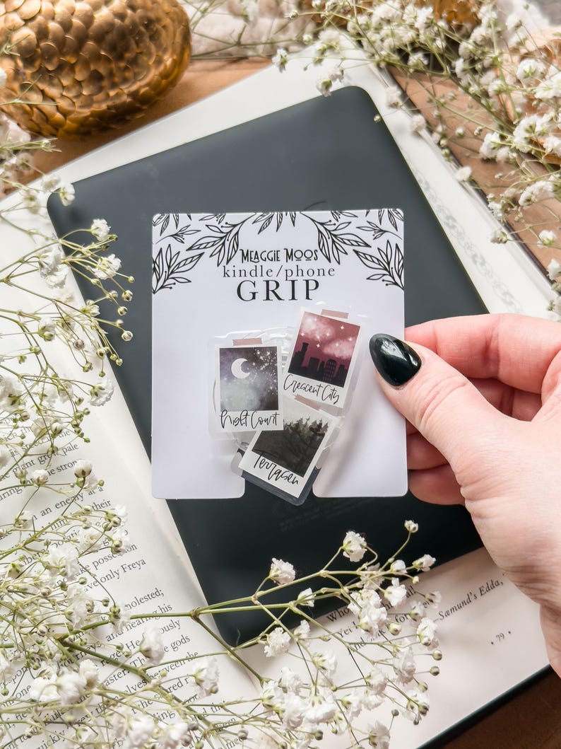 May include: A white card with the text "MEAGGIE MOOS kindle/phone GRIP" and three small, rectangular grips with landscape designs. The grips are held over a black e-reader and an open book with white flowers.