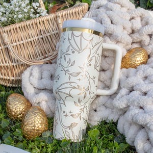 May include: A white stainless steel tumbler with a gold lid and a dragon design. The tumbler has a white handle and a straw. The tumbler is sitting on a white blanket with gold dragon eggs.