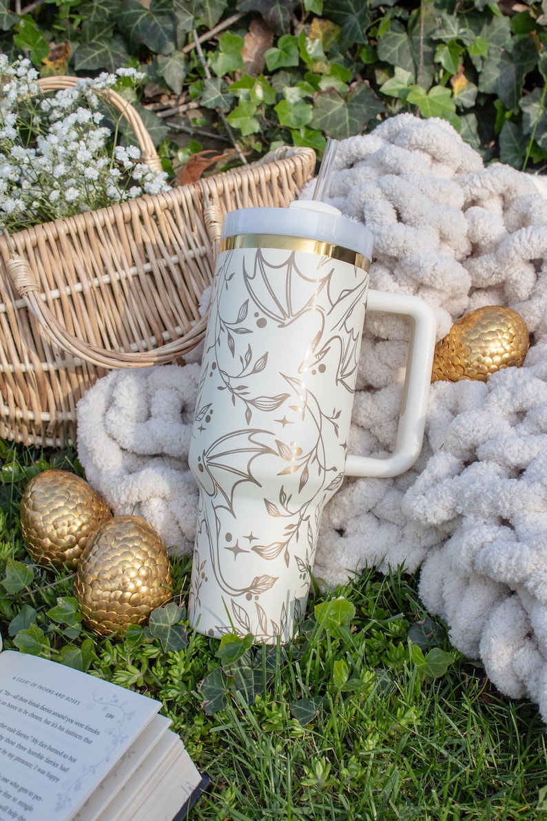 May include: A white stainless steel tumbler with a gold lid and a dragon design. The tumbler has a white handle and a straw. The tumbler is sitting on a white blanket with gold dragon eggs.