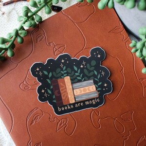 May include: A sticker with a stack of books, green leaves, and stars on a black background. The text "books are magic" is written in gold lettering below the books.