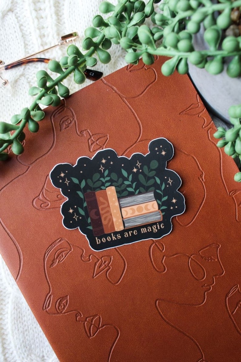May include: A sticker with a stack of books, green leaves, and stars on a black background. The text "books are magic" is written in gold lettering below the books.