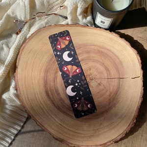 Puede incluir: Un marcap&aacute;ginas negro con un patr&oacute;n repetitivo de lunas blancas, estrellas doradas y polillas marrones con acentos rojos. El marcap&aacute;ginas est&aacute; sobre una superficie de madera.