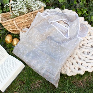 May include: A beige reusable shopping bag with a white floral pattern. The bag is folded and lying on a green grassy surface with a wicker basket, a book, and a white blanket.