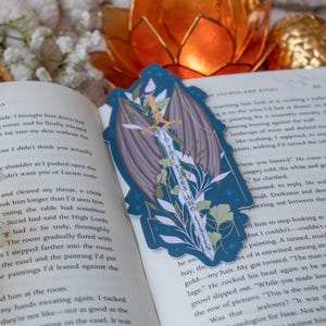 May include: A blue bookmark with a sword and wing design, with the text "THE STARS WHO LISTEN". The bookmark is placed on an open book with text. The background includes an orange flower and other decorative elements.