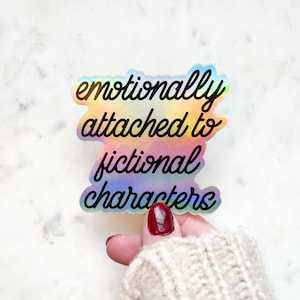 May include: A holographic sticker with the text "emotionally attached to fictional characters" in black lettering.