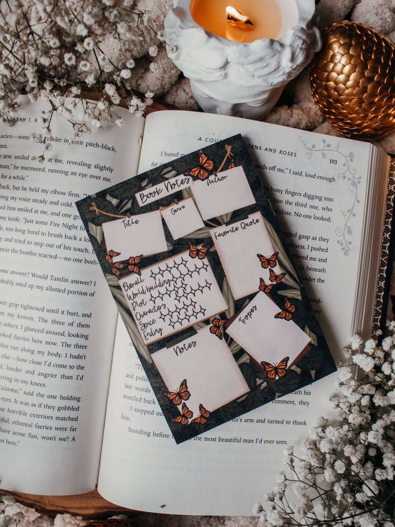 K&ouml;nnte beinhalten: Eine Notizkarte f&uuml;r B&uuml;cher mit dunkelgr&uuml;nem Hintergrund und orangefarbenen Schmetterlingen. Die Karte hat Abschnitte f&uuml;r Titel, Autor, Genre, Lieblingszitat und Notizen. Im Hintergrund ist ein Buch ge&ouml;ffnet.