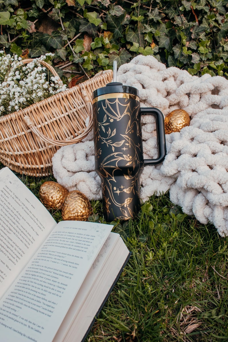 May include: A black and gold stainless steel tumbler with a dragon design. The tumbler has a lid and a handle. It is sitting on a bed of green grass with a book, a basket, and a white blanket.
