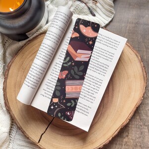 Puede incluir: Un marcapáginas decorativo con un diseño celestial, que incluye una luna creciente, estrellas y libros, insertado en un libro sobre una rodaja de madera. Una vela encendida y gafas están en el fondo. El marcapáginas tiene un fondo oscuro con elementos florales.