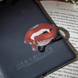 May include: A red and white vampire lips phone grip is attached to a black tablet. The lips have white fangs and a dripping effect. The tablet is resting on a book.