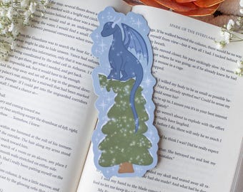 Könnte beinhalten: Blauer Drachen-Lesezeichen mit Baumdesign. Das Lesezeichen zeigt einen hellblauen Drachen, der auf einem grünen Baum sitzt, mit einem hellblauen Rand und Sternakzenten. Das Lesezeichen liegt auf einem offenen Buch.