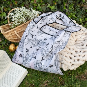 Peut inclure: Un sac de courses r&eacute;utilisable blanc avec un imprim&eacute; noir et blanc de motifs floraux et de chauves-souris. Le sac est pli&eacute; et pos&eacute; sur un lit d'herbe verte.