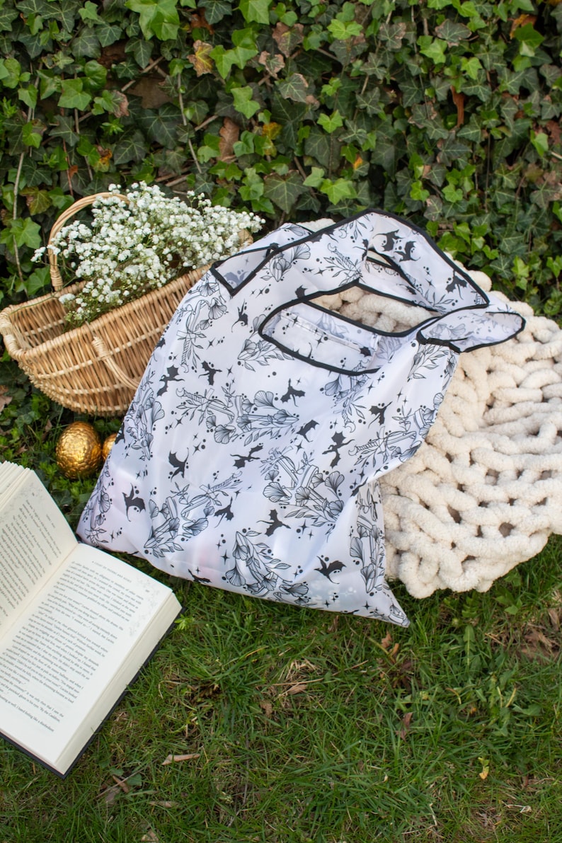 Peut inclure: Un sac de courses r&eacute;utilisable blanc avec un imprim&eacute; noir et blanc de motifs floraux et de chauves-souris. Le sac est pli&eacute; et pos&eacute; sur un lit d'herbe verte.