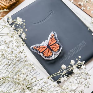 May include: A black e-reader with a monarch butterfly phone grip. The butterfly grip features orange and black wings. White flowers and a book are in the background.
