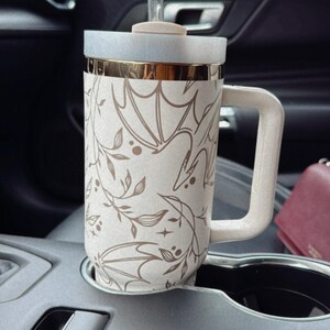 May include: A white and gold travel mug with a floral and leaf design. The mug has a lid and a straw. The mug is sitting in a cup holder in a car.