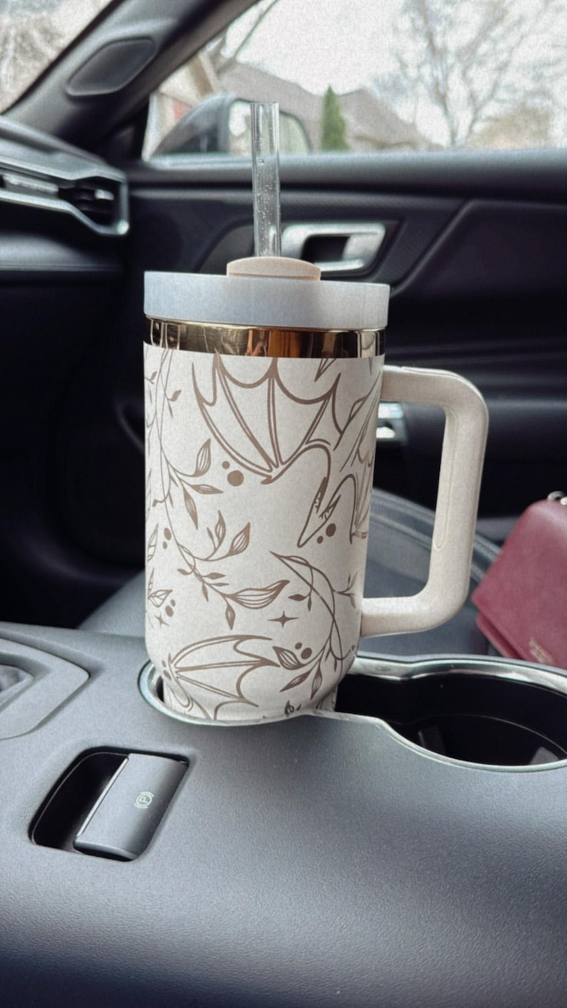 May include: A white and gold travel mug with a floral and leaf design. The mug has a lid and a straw. The mug is sitting in a cup holder in a car.