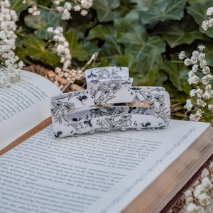 May include: A rectangular hair claw clip with a white base and a black line art pattern featuring various mushrooms, foliage, and small flying creatures. The clip has a hinged mechanism with interlocking teeth and a smooth, glossy finish.