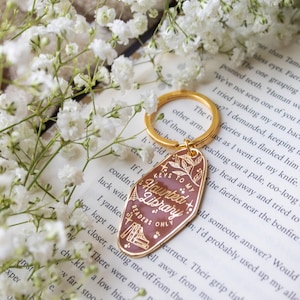 Puede incluir: Un llavero dorado con una etiqueta granate que dice "Keys to my Haunted Library Readers Only". El llavero est&aacute; rodeado de flores blancas y descansa sobre un libro.