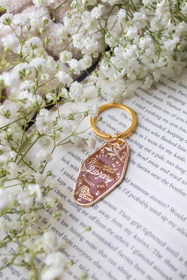 Puede incluir: Un llavero dorado con una etiqueta granate que dice "Keys to my Haunted Library Readers Only". El llavero est&aacute; rodeado de flores blancas y descansa sobre un libro.