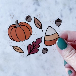 Puede incluir: Una pegatina transparente con una calabaza naranja, un caramelo de ma&iacute;z, bellotas marrones y hojas rojas y naranjas sobre un fondo blanco.