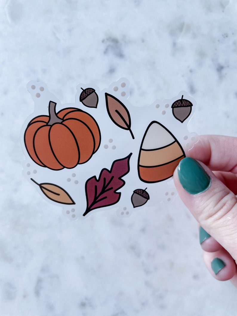 Puede incluir: Una pegatina transparente con una calabaza naranja, un caramelo de ma&iacute;z, bellotas marrones y hojas rojas y naranjas sobre un fondo blanco.