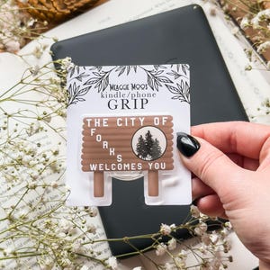 May include: A brown and white "The City of Forks Welcomes You" phone grip. The grip is in a package that says "MEAGGIE MOOS kindle/phone GRIP". The grip is on top of a black tablet and a book with white flowers.