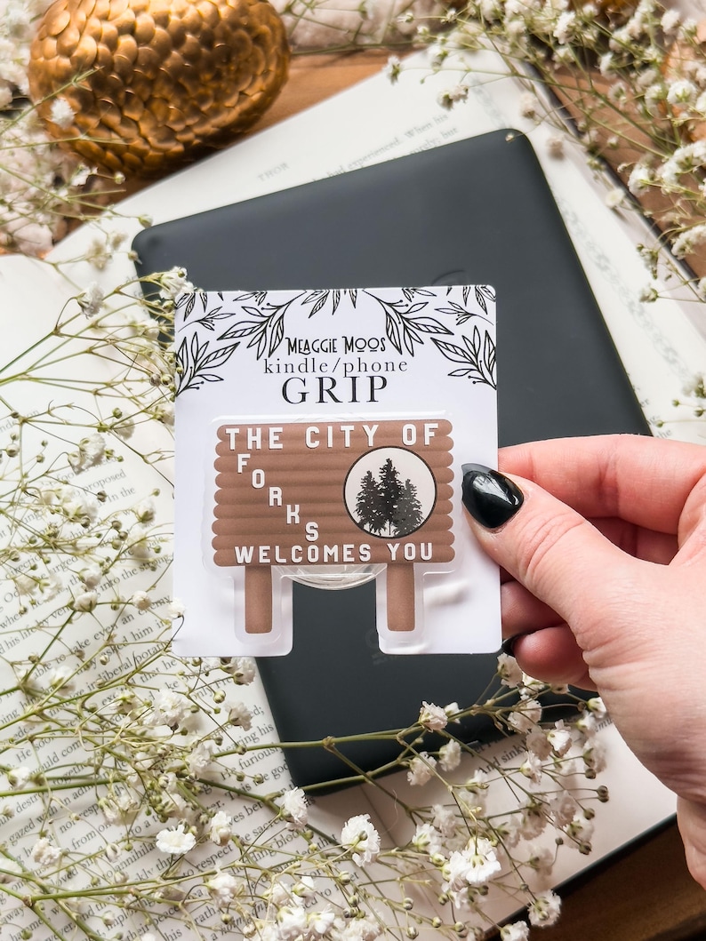 May include: A brown and white "The City of Forks Welcomes You" phone grip. The grip is in a package that says "MEAGGIE MOOS kindle/phone GRIP". The grip is on top of a black tablet and a book with white flowers.