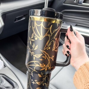 May include: A black and gold stainless steel travel mug with a black handle and a gold floral and bat design. The mug has a straw and is in a car cup holder.