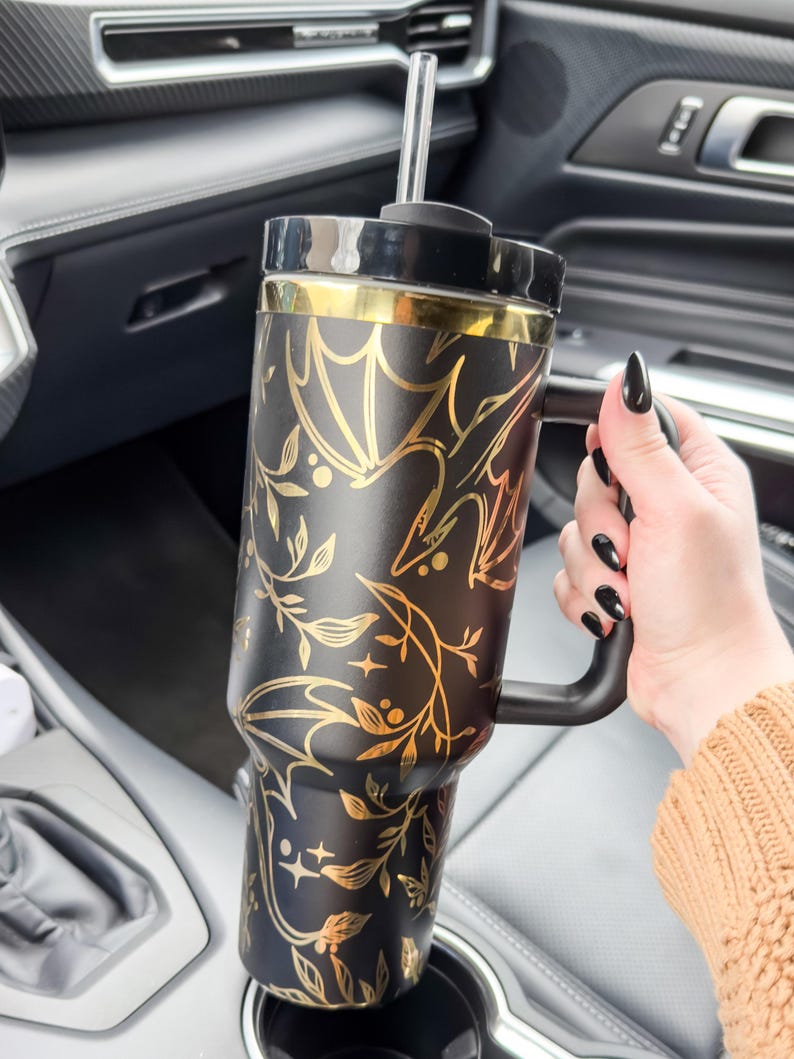 May include: A black and gold stainless steel travel mug with a black handle and a gold floral and bat design. The mug has a straw and is in a car cup holder.
