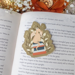 May include: A die-cut sticker featuring a light beige dragon perched atop a stack of three books, surrounded by stylized green foliage and small star accents. The sticker is placed on an open book page with visible printed text.