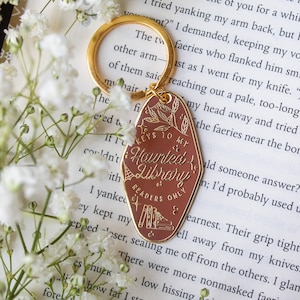 Puede incluir: Un llavero dorado con una etiqueta marr&oacute;n que dice "Keys to my Haunted Library & Readers Only". La etiqueta presenta un dise&ntilde;o bot&aacute;nico e ilustraciones de libros. El llavero est&aacute; sobre un libro y flores blancas.