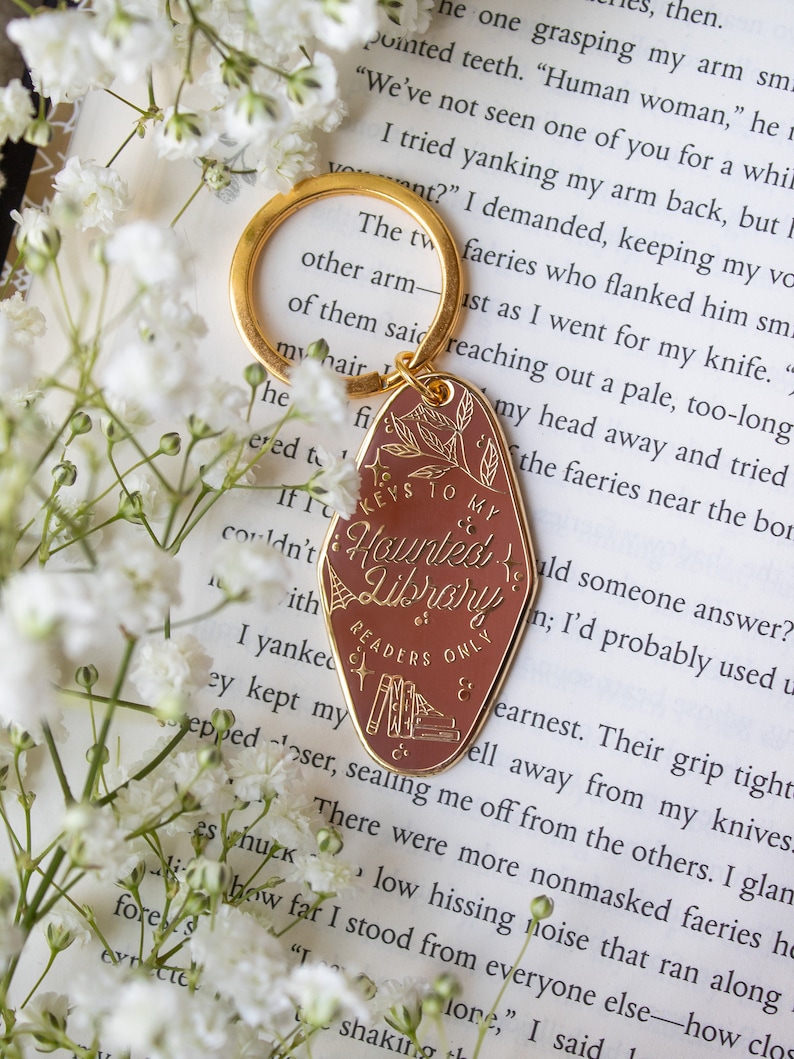 Puede incluir: Un llavero dorado con una etiqueta marr&oacute;n que dice "Keys to my Haunted Library & Readers Only". La etiqueta presenta un dise&ntilde;o bot&aacute;nico e ilustraciones de libros. El llavero est&aacute; sobre un libro y flores blancas.