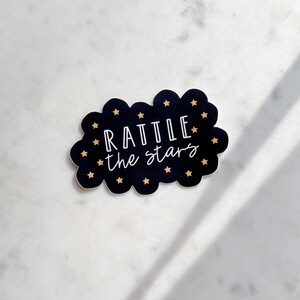 Peut inclure: Autocollant noir et blanc en forme de nuage avec des &eacute;toiles dor&eacute;es. Le texte "Rattle the stars" est &eacute;crit en blanc.