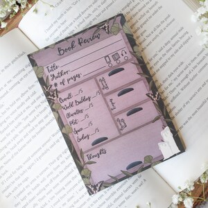 May include: A pink and green book review card with a floral design. The card has a section for rating the book on different aspects, such as world building, characters, plot, space, and ending. There is also a section for thoughts and a section for favorite tropes. The card is designed to look like a dresser with drawers.